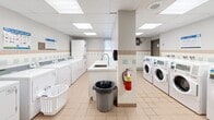 Located at 2645 Battleford Rd in Mississauga, Ontario, this shared laundry facility is a well-equipped, single-floor space designed for efficient and comfortable use. The room features a generous layout with multiple front-loading and top-loading washers and dryers arranged along the walls, offering ample capacity for residents. A central island with a utility sink provides a convenient space for hand-washing or pre-treating garments, while an adjacent marble-finish folding counter offers a clean, dedicated surface for sorting and folding laundry. A card-operated payment terminal is conveniently positioned for easy access. The space is finished with neutral ceramic tile flooring and a classic tile wainscoting with pastel accent tiles that run along the walls, lending a clean and functional aesthetic. A suspended ceiling with recessed panel lighting ensures bright, even illumination throughout. A window along one wall allows for natural light to filter in. Safety features include a visible fire extinguisher and a security camera. Rolling laundry carts and a seating bench are available for added convenience. Informational postings and laundry regulations are clearly displayed, making this facility straightforward and user-friendly.