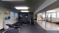 This single-floor fitness center is designed to accommodate a variety of workout needs within a functional, well-organized layout. The main exercise area features dark rubber flooring, which provides both durability and comfort underfoot during workouts. Cardio equipment is the focal point of the space, including a treadmill, stationary bike, and elliptical machine, all positioned to allow comfortable use without overcrowding.

A wall-mounted flat-screen television is installed above the equipment, offering entertainment during workouts. Motivational and informational posters are displayed on the deep blue-painted accent wall, adding visual interest and purpose to the space. Bright flush-mounted ceiling light panels illuminate the room evenly, ensuring a well-lit environment throughout.

A large mirror along one wall visually expands the space and allows users to monitor their form while exercising. Beyond the mirrored section, an adjacent area is visible, featuring lighter flooring and additional equipment, including what appears to be a punching bag, suggesting a dedicated strength or functional training zone. Windows along the side wall allow natural light to supplement the overhead lighting.