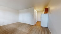 This compact yet functional 1-bedroom, 1-bathroom unit is thoughtfully laid out across a single floor, offering all the essentials in a straightforward, efficient design. The living room is a comfortable open space featuring neutral carpet flooring, white walls, and a sliding glass door with an adjacent window that brings in natural light and provides access to an outdoor balcony or patio area. A baseboard heating unit and wall-mounted air conditioning unit ensure year-round climate control. The kitchen is equipped with white cabinetry accented by warm wood trim, speckled laminate countertops, a stainless steel sink, a white electric range, and a full-size refrigerator. Upper cabinets offer ample storage space for everyday needs. A hallway connects the main living area to the bedroom and bathroom. The bedroom is a generously sized, carpeted room featuring a ceiling fan with a light fixture and a built-in closet with bifold doors and a full shelving and hanging rod system. The bathroom is fitted with a wood-trimmed vanity with a white countertop sink, a mirrored medicine cabinet for storage, and a bathtub. Hardwood-style flooring in the kitchen and hallway areas adds a practical contrast to the carpeted living and sleeping spaces throughout.