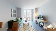 This single-floor residence showcases a thoughtfully designed interior with a cohesive contemporary aesthetic throughout. The living room serves as the centerpiece of the space, featuring dark hardwood flooring, soft gray walls, and floor-to-ceiling windows that frame sweeping urban skyline views and flood the room with natural light. A tufted emerald green sofa anchors the seating area, complemented by a geometric coffee table, a patterned area rug, and a gallery-style arrangement of framed mirrors. A dedicated media and workspace area with a sleek white console sits opposite, maximizing the room's functionality.

The kitchen is outfitted with light wood cabinetry, stainless steel appliances including a gas range and French door refrigerator, and a white quartz waterfall island with pendant lighting overhead, providing both prep space and casual seating.

The bedroom offers a tranquil retreat, distinguished by a bold dark accent wall, a large upholstered headboard, hardwood floors, and a window with city views. A well-appointed dresser with gold-accented décor adds warmth to the room.

The bathroom features large-format tile, a frameless glass walk-in shower, a modern vanity with a white quartz countertop, and clean chrome fixtures, completing this polished urban residence.