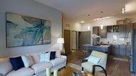 This single-floor residence offers a thoughtfully designed open-concept layout that connects the living area and kitchen in a cohesive, flowing arrangement. The living room is anchored by a light-toned sofa and complementary armchair, both accented with teal and navy throw pillows that echo the coastal-inspired abstract artwork adorning the soft grey accent wall. A glass-top coffee table and layered area rug add depth to the seating area, while warm wood-look flooring runs throughout, grounding the space with a natural, inviting feel.

The kitchen is visible from the living area and features dark cabinetry with sleek hardware, a light stone countertop, and a subway tile backsplash. Pendant lights hang above the kitchen bar, providing both task lighting and a refined decorative element. Track lighting overhead further enhances the kitchen's functionality.

A hallway visible in the background leads to additional rooms, suggesting a well-organized floor plan. The neutral color palette of warm beige, cool grey, and muted teal creates a calm, modern ambiance throughout, making this space feel both stylish and comfortable.