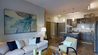 This single-floor residence offers a thoughtfully designed open-concept layout that connects the living area and kitchen in a cohesive, flowing arrangement. The living room is anchored by a light-toned sofa and complementary armchair, both accented with teal and navy throw pillows that echo the coastal-inspired abstract artwork adorning the soft grey accent wall. A glass-top coffee table and layered area rug add depth to the seating area, while warm wood-look flooring runs throughout, grounding the space with a natural, inviting feel.

The kitchen is visible from the living area and features dark cabinetry with sleek hardware, a light stone countertop, and a subway tile backsplash. Pendant lights hang above the kitchen bar, providing both task lighting and a refined decorative element. Track lighting overhead further enhances the kitchen's functionality.

A hallway visible in the background leads to additional rooms, suggesting a well-organized floor plan. The neutral color palette of warm beige, cool grey, and muted teal creates a calm, modern ambiance throughout, making this space feel both stylish and comfortable.