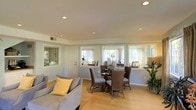 This single-floor home offers a warm and inviting atmosphere defined by clean lines and an open, airy layout. The combined living and dining area serves as the heart of the home, where light hardwood flooring flows seamlessly throughout, creating a cohesive and comfortable environment.

The dining area is centrally positioned and features a round glass-top table surrounded by woven rattan chairs, lending a relaxed yet refined character to the space. Adjacent to this, the living area provides a cozy retreat with a plush gray sofa accented by a soft yellow throw pillow, adding a touch of color to the neutral palette.

Natural light is a standout feature, with multiple windows dressed in sheer curtains lining the walls and allowing daylight to pour in from multiple directions. This creates a bright and cheerful ambiance throughout the day. Recessed ceiling lights provide additional illumination, ensuring the space remains well-lit at all times.

The walls are finished in a soft, neutral tone that complements the warm flooring and natural textures found in the furnishings. A lush potted plant near the corner adds a fresh, organic element to the interior. The overall design balances functionality and comfort, making this a welcoming and practical single-level home.