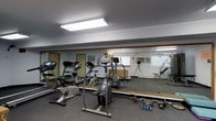 This single-floor fitness room offers a practical and well-organized workout space designed to accommodate a variety of exercise routines. The room is outfitted with an array of cardio equipment, including a treadmill, elliptical trainer, and stationary bike, giving users multiple options for cardiovascular exercise. A set of dumbbells and additional free weights are available for strength training, rounding out the room's functional fitness offerings. Wall-to-wall mirrors line one side of the space, visually expanding the room while allowing users to monitor their form during workouts. The flooring is covered in a durable, low-pile carpet suited for gym use. Overhead fluorescent lighting provides bright, even illumination throughout the space. A wall-mounted air conditioning unit ensures climate control for comfort during exercise. The neutral, light-colored walls contribute to a clean and focused atmosphere. A wooden bench or storage element is also visible, adding to the room's utility. Overall, this fitness room is a straightforward, no-frills space that prioritizes function and accessibility, making it well-suited as a shared or community-use gym facility.