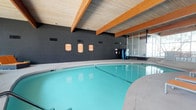 This indoor pool facility is a well-appointed aquatic space designed for both leisure and lap swimming. The pool itself is generously sized, featuring clean, clear turquoise water and a smooth surrounding deck that provides ample space for entry and exit. The architectural design is highlighted by a striking wood-paneled ceiling supported by exposed wooden beams, lending the space a warm and structured aesthetic that contrasts with the bold dark-painted walls. Large windows along one side of the room allow natural light to filter in, brightening the interior and creating an open, airy atmosphere despite the fully enclosed setting. Safety and informational signage is prominently displayed along the wall, ensuring guests are well-informed of pool rules and guidelines. A water vending station is conveniently positioned near the pool deck, adding a practical amenity for swimmers. The overall ambiance is clean, functional, and inviting, making this indoor pool a capable facility suited for year-round aquatic use regardless of outdoor weather conditions.