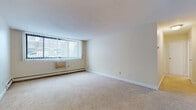 This one-bedroom, one-bathroom unit is located at 3029 France Ave S in Minneapolis, MN, offering a practical and comfortable single-floor layout. The living area is generously sized, featuring neutral wall-to-wall carpeting, soft cream-toned walls, and large windows with vertical blinds that allow natural light to fill the space. A baseboard heating system and a wall-mounted AC unit ensure year-round climate control. The bedroom mirrors the living area's carpeted flooring and light-filled atmosphere, with its own large window and baseboard heating, providing a quiet and comfortable retreat. The kitchen is efficiently designed with dark wood cabinetry, a gas range, a refrigerator, and a tile backsplash, opening conveniently toward a hallway that connects the main living spaces. The full bathroom features classic white tile walls and a mosaic tile floor with accent detailing, complemented by a tub-shower combination, a white vanity with cabinet storage, and a medicine cabinet mirror. Throughout the unit, clean white trim and textured ceilings contribute to a cohesive, understated aesthetic. The overall layout is functional and straightforward, making efficient use of the available space.