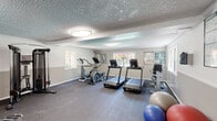Located at 10453 Artesia Boulevard in Bellflower, California, this fitness center offers a well-organized workout facility designed to support a range of exercise needs. The main workout area features durable rubber flooring throughout, providing a safe and functional surface for both cardio and strength training activities. Multiple treadmills and an elliptical machine are arranged along the windowed walls, where several windows allow natural light to fill the space and offer views of the surrounding outdoor area. A cable resistance machine and a dumbbell rack with a full set of weights cater to strength training, while a weight bench adds versatility for various exercises. Exercise balls are available in the corner for additional fitness options. Floor-to-ceiling mirrors line one wall, enhancing the sense of space and allowing users to monitor their form during workouts. Informational fitness posters are displayed on the walls for guidance and reference. The space is illuminated by flush-mount ceiling light fixtures set against a textured ceiling, creating a bright and functional atmosphere. A wall-mounted air conditioning unit ensures climate comfort year-round. An adjacent restroom with tile flooring, a toilet, and a sink provides convenient facilities for gym users.