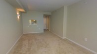 This single-floor residential unit at 1310 offers a straightforward and functional layout designed for comfortable everyday living. The main living area is generously proportioned, featuring neutral-toned wall-to-wall carpeting that adds warmth underfoot and light sage-green walls that create a calm, understated ambiance throughout the space. A notable architectural detail is the pass-through window connecting the living area to the kitchen, which enhances the sense of openness and allows for easy interaction between the two spaces. The kitchen is equipped with wood-toned cabinetry and includes a refrigerator and range, providing all the essentials in a compact, efficient layout. The entryway, illuminated by overhead lighting, features a tiled floor and a solid door that opens to the unit, offering a clean and welcoming transition into the home. White baseboards throughout provide a crisp contrast against the carpeted floors and painted walls, contributing to a tidy, well-maintained appearance. The textured ceiling adds a subtle design element consistent across the living spaces. Overall, the unit presents a clean, move-in-ready interior with a cohesive neutral palette that offers a versatile canvas for personalization.