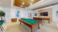 Located at 17475 S Dixie Hwy in Palmetto Bay, Florida, this single-floor recreational and lounge space is thoughtfully designed to blend entertainment with contemporary style. The room is anchored by a full-size pool table and a foosball table, making it an ideal gathering spot for social activity. The ceiling is a standout architectural feature, showcasing warm wood paneling set within a coffered design with recessed and track lighting that creates a welcoming, well-lit atmosphere throughout.

The flooring consists of large-format light-toned tiles that contribute to a clean, modern aesthetic. Distinct zones within the space serve different purposes: a lounge corner features tufted caramel leather sofas paired with a gold pedestal coffee table set with a chess board, all beneath large-scale abstract artwork in blue and teal tones. A bar-height bistro table with teal chairs is positioned near floor-to-ceiling windows draped with sheer curtains, offering views of an outdoor pool area and filling the space with natural light.

An accent wall clad in glossy emerald green tile frames a mounted flat-screen television alongside built-in cabinetry, adding a bold pop of color. Lush potted plants and curated artwork throughout complete the vibrant, resort-inspired ambiance of this space.