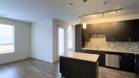 This single-floor space showcases a sleek, modern design centered around an open-concept layout that seamlessly connects the kitchen and living areas. The kitchen is a standout feature, highlighted by rich dark cabinetry that extends to the ceiling, offering generous storage. A central island with a light-toned granite countertop serves as both a workspace and a casual gathering point, anchored by a bold dark base that contrasts elegantly with the stone surface. Pendant lighting hangs above the island, while a track lighting system overhead ensures the kitchen remains well-lit throughout. A multicolored mosaic tile backsplash adds texture and visual interest between the cabinetry, complementing the overall contemporary aesthetic. Stainless steel appliances contribute to the polished, professional feel of the cooking space. The flooring throughout is a warm-toned hardwood-style material that runs continuously, unifying the open space. Natural light filters in through a window fitted with horizontal blinds, brightening the adjacent living area. The neutral wall tones and clean lines throughout create a cohesive, inviting atmosphere well-suited to modern living.