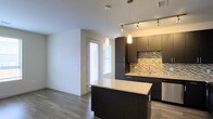 This single-floor space showcases a sleek, modern design centered around an open-concept layout that seamlessly connects the kitchen and living areas. The kitchen is a standout feature, highlighted by rich dark cabinetry that extends to the ceiling, offering generous storage. A central island with a light-toned granite countertop serves as both a workspace and a casual gathering point, anchored by a bold dark base that contrasts elegantly with the stone surface. Pendant lighting hangs above the island, while a track lighting system overhead ensures the kitchen remains well-lit throughout. A multicolored mosaic tile backsplash adds texture and visual interest between the cabinetry, complementing the overall contemporary aesthetic. Stainless steel appliances contribute to the polished, professional feel of the cooking space. The flooring throughout is a warm-toned hardwood-style material that runs continuously, unifying the open space. Natural light filters in through a window fitted with horizontal blinds, brightening the adjacent living area. The neutral wall tones and clean lines throughout create a cohesive, inviting atmosphere well-suited to modern living.