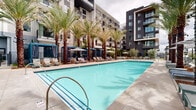 This expansive outdoor pool area at Alexan Camellia in San Diego, CA offers a resort-caliber amenity experience nestled within a modern residential community. At the heart of the space is a generously sized rectangular swimming pool with vibrant turquoise water, providing an inviting focal point for relaxation and recreation. The pool deck is thoughtfully designed with a wide surrounding walkway that accommodates multiple rows of cushioned lounge chairs and chaise seating, ideal for soaking up the Southern California sun. Along one side of the pool, a series of elegant private cabanas with teal and white drapery offer shaded retreats, each furnished for comfort and a touch of luxury. Striped market umbrellas are strategically positioned throughout the deck, providing additional shade options. Towering mature palm trees are planted throughout the courtyard, lending a lush, tropical ambiance and framing views of the surrounding multi-story residential buildings. Lush low-level greenery and potted plants add to the landscaped aesthetic. The overall design balances open, sun-drenched areas with shaded lounging zones, creating a versatile outdoor retreat that captures the relaxed, upscale spirit of San Diego living.
