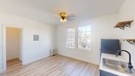 This well-appointed single-floor space offers a clean, modern aesthetic complemented by classic architectural details. The main living area is filled with natural light streaming through large, multi-pane windows that frame views of the surrounding urban streetscape. Light hardwood flooring runs throughout, creating a warm and cohesive feel across all rooms. A ceiling fan with a gold-toned fixture adds a touch of vintage charm to the living space, while a cast-iron radiator provides efficient heating. The kitchen area features navy blue cabinetry, adding a bold accent against the otherwise crisp white walls and trim. Crown molding details elevate the interior with a refined finish. A generously sized closet with built-in shelving offers practical storage, while additional doorways lead to a separate hallway and utility areas. The bathroom is a standout feature, showcasing floor-to-ceiling white subway tile with dark grout, matte black fixtures, and a combination tub and shower. A window above the tub brings in natural light and opens to an outdoor view, enhancing the airy feel. Throughout the space, clean white walls, modern hardware, and thoughtful finishes create a bright, move-in-ready environment with a cohesive and contemporary character.