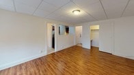 This single-floor space offers a practical and well-organized layout with a variety of functional areas flowing seamlessly from one to the next. The main living area is bright and open, featuring warm hardwood-style flooring that runs throughout, crisp white walls, and a drop tile ceiling with flush lighting. Two windows along one wall allow natural light to fill the room, creating an airy and welcoming atmosphere. A radiator beneath the windows provides efficient heating. The living space connects to additional rooms via clean white-trimmed doorways, one of which reveals a closet with built-in shelving, offering useful storage.

The kitchen is compact yet well-appointed, showcasing rich dark cherry wood cabinetry with raised-panel detailing, granite countertops with warm gold and brown tones, and a stainless steel electric range with an overhead ventilation hood. The combination of quality materials gives the kitchen a polished, updated feel.

The bathroom continues the classic aesthetic with sage green ceramic tile wainscoting, small white mosaic floor tiles, and a white pedestal-style sink. A frosted window provides natural light while maintaining privacy. The bathtub and shower area is fully tiled, and a wall-mounted mirror and vanity lighting complete the functional space. Overall, the unit presents a clean, move-in ready environment with character and practicality throughout.