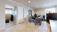 This single-story leasing office is designed to support both staff operations and resident or prospective tenant interactions. The space is finished with large-format beige ceramic tile flooring and crown molding throughout, lending a clean, professional appearance.

The reception area features a wood-framed front desk with open shelving and a light countertop, accompanied by a wall-mounted flat-screen television — useful for displaying community information. Adjacent to this area, a private staff workspace is equipped with dark wood desks, computer workstations, and office chairs, providing a functional environment for leasing personnel.

A dedicated consultation area includes a wooden meeting table flanked by upholstered armchairs, ideal for one-on-one discussions with prospective residents. Framed artwork and colorful wall décor contribute to a welcoming ambiance. Recessed lighting illuminates the meeting area, while natural light filters in through a windowed exterior door.

The office also includes a compact kitchen area visible in the layout, and interior doors provide access to additional private spaces. Overall, the leasing office balances practicality with an approachable, well-maintained aesthetic suited for a professional residential community setting.