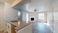 This single-story unit is designed with a functional, open-concept layout that seamlessly connects the kitchen and living areas. The kitchen is well-appointed with white shaker-style cabinetry, a white subway tile backsplash, stainless steel appliances including a refrigerator, electric range, and microwave, and a central island with a dark quartz countertop and integrated sink. A built-in shelving unit adjacent to the kitchen provides additional storage.

The living area is bright and inviting, benefiting from multiple windows fitted with blinds that allow ample natural light to fill the space. A corner fireplace with a white surround and brick detailing serves as a focal point, while a ceiling fan overhead adds comfort. Wood-style plank flooring extends throughout the main living spaces and bedroom, contributing to a cohesive and modern aesthetic.

The bedroom is comfortably sized, featuring a ceiling fan, a large window for natural light, and direct access to a bathroom. The bathroom showcases a floating vanity with a walnut-toned cabinet, a white integrated sink, and a framed mirror. A dedicated laundry area houses a full-sized top-load washer and dryer with overhead shelving for added convenience. Textured ceilings are present throughout the unit.