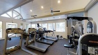 This fitness center is designed to support a well-rounded workout experience within a single-level space. The room is outfitted with multiple treadmills and elliptical machines positioned along the windowed wall, allowing users to exercise while enjoying views of the surrounding landscape. Natural light streams in through a row of large windows, creating a bright and energizing atmosphere throughout the space.

The flooring is covered in durable rubber gym matting, providing both safety and comfort underfoot. Ceiling fans are mounted above to aid in air circulation and maintain a comfortable temperature during workouts. Recessed lighting supplements the natural light, ensuring the space remains well-lit at all times of day.

Strength training is accommodated with a cable machine and additional resistance equipment positioned to one side of the room. A floor-to-ceiling mirror lines one wall, enhancing the sense of space and allowing users to monitor their form during exercise. The overall layout is thoughtfully organized to maximize usability, offering a clean, functional environment suitable for a variety of fitness routines.