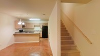Located at 26676 Canal Rd. in Orange Beach, Alabama, this two-story property offers a functional and straightforward layout spread across two levels. The main floor showcases an open kitchen area equipped with light wood cabinetry, a built-in cooktop, a sink with surrounding counter space, and a full-size refrigerator. A kitchen island provides additional workspace and adds to the open feel of the area. The flooring on the main level features a warm-toned concrete surface, lending a durable and easy-to-maintain finish to the space. Overhead fluorescent lighting illuminates the kitchen area efficiently. A staircase with a wood handrail leads from the main floor to the upper level, offering access to the additional living areas above. The interior palette is kept neutral throughout, with light-colored walls that contribute to a clean and open atmosphere. The overall design emphasizes practicality and ease of use, making this Orange Beach property a functional space suited for a variety of uses.
