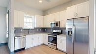 This thoughtfully designed single-floor residence combines contemporary finishes with a clean, cohesive aesthetic throughout. The kitchen serves as a functional centerpiece, featuring white shaker-style cabinetry extending to the ceiling for ample storage, granite countertops, a gray subway tile backsplash, and a full suite of stainless steel appliances including a French door refrigerator with water dispenser, electric range, over-the-range microwave, and dishwasher. Light streams in through a window above the sink, brightening the space naturally.

The living and entry areas showcase light-toned luxury vinyl plank flooring and neutral gray walls, unified by striking teal-blue accent doors and bold matching baseboards that add a distinctive design touch throughout. French doors in the main living area invite natural light and provide outdoor access.

The bedrooms offer plush carpeting underfoot, generous windows with blinds for natural light and privacy, and closet access through the signature blue panel doors. A hallway connects the rooms seamlessly, maintaining an open, organized flow.

The bathroom is outfitted with granite countertops, a white undermount sink, a large framed mirror, and a tiled shower-tub combination, all set above matching vinyl flooring. Every detail reflects a modern, move-in-ready finish.