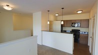 This single-floor space showcases an open-concept design that integrates the kitchen and living area into one cohesive environment. The kitchen is a functional centerpiece, featuring a white peninsula with dark countertops that provides both additional workspace and a natural dividing point between the cooking and living zones. Medium-toned cabinetry lines the kitchen walls, complemented by black appliances including a range and microwave, creating a classic, contrasting aesthetic. Two elegant pendant lights hang above the peninsula, adding focused task lighting while enhancing the overall visual appeal of the space. A flush-mount ceiling fixture provides additional ambient lighting in the kitchen area. The transition from the kitchen's hard flooring to the plush carpeting of the adjacent living space adds textural variety while reinforcing the distinct yet connected nature of each zone. White walls and trim throughout the space lend a clean, neutral backdrop that allows the darker kitchen elements to stand out. The open layout promotes an easy flow between cooking, dining, and living areas, making the space well-suited for both everyday living and casual entertaining.