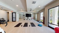 This single-floor amenity space is thoughtfully designed to offer both entertainment and relaxation in a cohesive, coastal-inspired aesthetic. The layout flows between two primary zones: an expansive game room and an open lounge area, connected by wide, light-toned hardwood flooring that runs throughout.

The game room is generously proportioned, anchored by a blue felt billiards table with a substantial pedestal base and complemented by a foosball table and a shuffleboard table. Woven pendant lights hang overhead from a shiplap ceiling, lending warmth and texture to the space. A wall-mounted flat-screen television is framed within a built-in white surround, while a curved banquette seating nook — upholstered in soft blue fabric and adorned with decorative woven wall art — provides an intimate gathering spot. A cue rack and small bistro tables complete the room's functional offerings.

The adjacent lounge area commands attention with dramatic double-height ceilings clad in botanical-patterned wallcovering. Large windows flood the space with natural light, highlighting the neutral furnishings, layered textiles, and a mix of seating that includes sofas, accent chairs, and woven ottomans. A rattan credenza and curated artwork add refined finishing touches throughout.