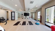 This single-floor amenity space is thoughtfully designed to offer both entertainment and relaxation in a cohesive, coastal-inspired aesthetic. The layout flows between two primary zones: an expansive game room and an open lounge area, connected by wide, light-toned hardwood flooring that runs throughout.

The game room is generously proportioned, anchored by a blue felt billiards table with a substantial pedestal base and complemented by a foosball table and a shuffleboard table. Woven pendant lights hang overhead from a shiplap ceiling, lending warmth and texture to the space. A wall-mounted flat-screen television is framed within a built-in white surround, while a curved banquette seating nook — upholstered in soft blue fabric and adorned with decorative woven wall art — provides an intimate gathering spot. A cue rack and small bistro tables complete the room's functional offerings.

The adjacent lounge area commands attention with dramatic double-height ceilings clad in botanical-patterned wallcovering. Large windows flood the space with natural light, highlighting the neutral furnishings, layered textiles, and a mix of seating that includes sofas, accent chairs, and woven ottomans. A rattan credenza and curated artwork add refined finishing touches throughout.