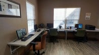 This single-floor office space is designed for productivity and collaboration, offering a well-organized and comfortable working environment. The room features multiple workstations arranged along the walls, each equipped with desktop computers, keyboards, and monitors, providing dedicated setups for individual users. The desks are finished in a clean, light tone, complementing the warm, neutral-colored walls that give the space a professional yet welcoming ambiance. Ergonomic office chairs in earthy tones of orange and green are positioned at each station, adding a touch of color to the room. The flooring consists of wood-look planks in a medium brown tone, lending warmth and a polished aesthetic underfoot. Two large windows fitted with horizontal blinds allow generous natural light to filter into the space, creating a bright and pleasant atmosphere while offering adjustable light control throughout the day. Small potted plants are placed on the desks, introducing subtle greenery that enhances the overall work environment. The layout is efficient and thoughtfully arranged, making good use of the available space to accommodate multiple users comfortably within a single, cohesive office setting.