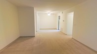 This single-floor space presents an open and functional layout finished in soft, neutral tones throughout. The main living area is generously sized, featuring wall-to-wall beige carpeting that lends a warm, cohesive feel underfoot. Smooth white walls and a light-colored ceiling create a bright, airy atmosphere, while a ceiling fan with a built-in light fixture serves as a practical central fixture overhead.

The layout flows openly from one area to the next, with a wide passageway connecting the main living space to adjacent rooms. Arched and rectangular doorways frame transitions between spaces, adding subtle architectural character to an otherwise straightforward design. A thermostat is visibly mounted on one wall, and a floor-level vent indicates a standard forced-air climate control system.

The space benefits from a clean, uncluttered feel, offering a versatile blank canvas suitable for a variety of furniture arrangements and uses. Natural and artificial light work together to keep the interior feeling open. Overall, the unit presents a practical, well-maintained environment with a simple, livable aesthetic.