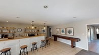 This single-floor space is thoughtfully designed as an entertainment and recreation area, combining a functional bar setup with a game area to create a cohesive, leisure-focused environment. The bar section features a white paneled base with a warm butcher block-style countertop, open wooden shelving above, and pendant lighting that casts a welcoming glow over the workspace. A row of industrial-style adjustable bar stools lines the counter, providing comfortable seating for guests. The open shelving displays decorative items and barware, adding a personalized touch to the space.

Adjacent to the bar, the recreation area centers on a full-length wooden shuffleboard table, a standout feature that anchors the room. Three framed botanical prints in blue and beige tones hang above the shuffleboard table, reinforcing a coastal aesthetic that carries throughout the space. The walls are finished in a soft neutral tone, complementing the light beige tile flooring that runs continuously across the room, contributing to a clean and cohesive look. Recessed lighting supplements the pendant fixtures, ensuring the space is well-lit throughout. A textured ceiling adds subtle visual interest overhead. The overall ambiance is casual yet polished, making it well-suited for both entertaining and everyday use.