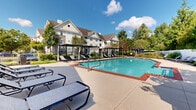 Located at 1515 Manley Rd in West Chester, PA, this outdoor amenity area centers around a generously sized swimming pool with crystal-clear blue water and brick-trimmed edges. The pool deck is finished in smooth concrete and outfitted with numerous lounge chairs, offering ample space for sunbathing and relaxation. A shallow wading section provides a gentle entry point into the pool, making it accessible and family-friendly.

Surrounding the pool are several sleek, modern pergola-style cabanas that provide shaded seating areas, adding both function and a resort-like aesthetic to the space. The perimeter is enclosed by a wrought iron fence, ensuring privacy and safety, while mature trees and well-maintained landscaping create a lush, green backdrop that enhances the tranquil ambiance.

A clubhouse-style building with large multi-pane windows and a classic gabled roofline sits adjacent to the pool area, offering additional facilities and views of the outdoor space. On the far end of the complex, a gazebo and additional seating areas provide a quieter retreat surrounded by greenery. The overall setting, framed by the multi-story residential buildings of the community, reflects a well-appointed outdoor recreational space designed for comfort and leisure in a serene suburban environment.