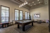 This single-floor community amenity space is thoughtfully designed to offer both social and functional areas for residents. The layout flows between distinct zones, each serving a unique purpose while maintaining a cohesive modern aesthetic throughout.

The lounge area is anchored by a tufted gray sofa, leather accent chairs, and a round white coffee table, creating an inviting setting for relaxation and conversation. Large windows with horizontal blinds flood the space with natural light and provide views of lush greenery outside. A bold, large-scale abstract artwork serves as a striking focal point on the wall, complemented by tall indoor plants and a brass floor lamp that add warmth and texture.

The game room zone features a full-size billiards table positioned beneath contemporary pendant lighting, with a wall-mounted flat-screen TV nearby. Additional high-top seating provides space for spectators or casual gatherings. A dedicated computer workstation area lines one wall, equipped with multiple desktop computers and a printer, offering a practical business center within the amenity space.

Architecturally, the space benefits from notably high ceilings with coffered and beamed detailing, arched doorways, and a neutral color palette of warm grays and beige tones. Carpeted flooring throughout adds comfort underfoot, while the open, column-accented layout contributes to an airy and expansive atmosphere.