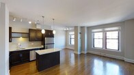 This well-appointed single-floor residence seamlessly blends the kitchen and living areas into one cohesive, open-plan layout. The kitchen is a standout feature, showcasing rich dark cabinetry, granite countertops, stainless steel appliances including a refrigerator and dishwasher, and a central island that serves as both a workspace and casual gathering spot. Elegant pendant lighting hangs above the island, adding warmth and a modern aesthetic. Track lighting runs throughout, providing flexible, layered illumination across the space. Gleaming hardwood floors extend throughout the entire area, contributing to a sense of continuity and warmth. The living area is generously proportioned, benefiting from multiple large windows that invite abundant natural light and frame views of the surrounding environment. The neutral gray walls and crisp white ceilings create a clean, versatile backdrop that complements the dark wood tones of the kitchen fixtures. Architectural details such as wide baseboards and structured column accents add a polished, urban character to the overall design. The open flow between the kitchen and living space makes this layout ideal for both everyday living and entertaining.