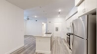 This single-floor residence presents a clean, contemporary design characterized by an all-white color palette, light wood-style luxury vinyl plank flooring, and recessed lighting throughout. The open-concept kitchen serves as the centerpiece of the living area, anchored by a central island with a granite countertop and an integrated double-basin sink. Crisp white cabinetry extends to the ceiling, providing ample storage, while a full suite of stainless steel appliances — including a gas range, over-the-range microwave, and French door refrigerator — lends a modern, functional aesthetic. Open shelving along one kitchen wall adds additional storage versatility.

The living area flows naturally from the kitchen and is equipped with a ceiling fan and recessed lighting. Adjacent rooms offer flexible living spaces, each finished with matching flooring and large windows fitted with white blinds that allow generous natural light. The bedrooms are well-proportioned, with ceiling fixtures and direct access to the main corridor. A dedicated laundry area houses a full-size washer and dryer side by side. Throughout the unit, clean white walls, consistent flooring, and streamlined fixtures contribute to a cohesive, move-in-ready atmosphere.