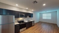This single-floor space showcases a clean, contemporary design with a thoughtful layout and quality finishes throughout. The open-concept kitchen and living area serves as the heart of the unit, featuring rich dark cabinetry, crisp white countertops, and a full suite of stainless steel appliances including a refrigerator, electric range, over-the-range microwave, and dishwasher. Warm-toned hardwood flooring extends across the kitchen and living area, lending a welcoming and cohesive feel to the space.

The bedrooms offer a comfortable contrast with plush wall-to-wall carpeting and neutral walls that create a calm, restful atmosphere. Natural light filters in through windows fitted with horizontal blinds. At least one bedroom includes a built-in closet with a shelving system, providing practical storage solutions.

The bathroom is well-appointed with a tub and shower combination, a sleek dark-toned vanity with a white countertop, an oval mirror, and a bright vanity light fixture above — all set against light-colored walls and hardwood-style flooring. Recessed lighting throughout the unit ensures even, modern illumination. Overall, the space presents a fresh, move-in-ready environment defined by its modern aesthetic and functional design.