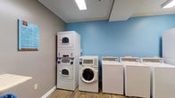 This communal laundry facility is a practical and well-maintained space designed to accommodate multiple users efficiently. The room is finished with a calming two-tone color scheme, featuring soft blue accent walls complemented by neutral gray-painted walls, lending the space a clean and welcoming atmosphere. Crown molding adds a refined touch to the interior, while wood-look flooring provides a durable and easy-to-clean surface underfoot. Overhead, flush-mounted fluorescent lighting ensures the room is brightly and evenly illuminated throughout.

The laundry room is outfitted with a comprehensive selection of commercial-grade appliances, including front-loading washing machines, top-loading washers, and stacked washer-dryer units — offering flexibility to suit varying laundry needs. The machines are arranged thoughtfully along the walls to maximize the usable floor space. Each unit features digital controls, reflecting a modern and user-friendly design. White baseboards frame the perimeter of the room, contributing to the overall crisp and tidy aesthetic. The space is single-floor and efficiently laid out, making it easy to navigate and use comfortably.