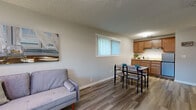 This thoughtfully laid-out studio occupies a single floor and makes efficient use of its compact footprint. The open-plan living area serves as the central hub of the space, featuring warm yellow-toned walls, a textured popcorn ceiling, and light wood-look laminate flooring that runs throughout. A large window along the exterior wall allows natural light to fill the room while offering views of surrounding greenery. A wall-mounted PTAC unit provides climate control directly beneath the window.

The living zone accommodates both a seating area and a dining space, with the open layout allowing flexible arrangement. A partial-height kitchen counter separates the kitchenette from the main living area, where warm wood cabinetry, a range hood, and a full-size refrigerator are visible. The sleeping area is integrated into the main open space, accessible via the hallway, which also connects to the bathroom and closet.

The bathroom is well-appointed with warm wood cabinetry, granite-style countertops, an undermount sink, and a glass-door shower enclosure, all set on matching wood-look flooring. Overall, the space presents a functional and cohesive design suited for single-occupancy living.