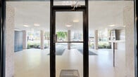 Located at 1655 Haro Street in Vancouver's West End neighbourhood, this building entrance hallway provides a clean and welcoming transition between the outdoors and the building's interior. The entry sequence begins with a covered exterior vestibule supported by textured concrete columns, where large glass doors display the building's address prominently above and open onto a "Welcome" mat leading inside.

The interior hallway is finished to a high standard, featuring large-format light beige ceramic tile flooring that extends throughout the space. One wall is clad in elegant marble-look panels, adding a refined, modern character to the corridor. A striking framed display case set into the wall houses a curated arrangement of tropical greenery, including a peace lily and succulents, serving as a decorative focal point that brings a touch of nature indoors.

Illumination is provided by flush-mounted ceiling light fixtures complemented by a wall-mounted lantern-style sconce, creating a warm and well-lit environment. The hallway leads to resident unit doors as well as elevator access, with an exit sign marking the corridor's connection to the broader building circulation. The overall ambiance is tidy, bright, and professionally maintained, reflecting a well-kept residential building in one of Vancouver's most desirable neighbourhoods.
