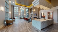 This elegantly designed clubhouse lounge is a standout amenity space that impresses from the moment you enter. The centerpiece of the main lounge is its dramatic double-height ceiling, accentuated by a gold-trimmed tray detail and a cascading modern chandelier with sculptural ring fixtures that add a contemporary artistic flair. Wide-plank wood-look flooring runs throughout, complementing the warm, neutral tones of the walls and furnishings.

Floor-to-ceiling grid windows line multiple walls, flooding the interior with abundant natural light and offering views of the surrounding outdoor pool area and landscaping. The main seating area is anchored by a sleek linear fireplace set into a textured stone accent wall, with a mounted flat-screen television above. Comfortable sectional sofas and accent chairs in teal, cream, and rust tones create inviting conversation areas.

Adjacent to the lounge is a bar and counter area featuring a bold geometric tile backsplash, bar seating, and stainless steel appliances. A large marble-topped island with white cabinetry anchors the entry area, ideal for gatherings or events. Intimate dining nooks positioned near the tall windows offer cozy spots bathed in natural light.

A dedicated private workstation room provides a quiet retreat, complete with a built-in white desk, computer setup, and stylish décor, making it a functional space for productivity within this beautifully appointed communal environment.