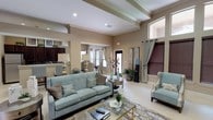 Located at 4500 Highway 57 in Ocean Springs, Mississippi, this single-story community space offers a refined and welcoming environment designed for both relaxation and social interaction. The main lounge area is anchored by a plush teal sofa and coordinating accent chairs arranged around a glass-top coffee table, all set atop a decorative area rug. Soaring ceilings with elegant tray detailing and crown molding add architectural depth, while recessed lighting and large windows allow natural light to fill the room with a warm, airy ambiance. A standout fireplace with a classic white surround serves as a focal point in an adjoining sitting area, complemented by a mounted flat-screen television and tall windows fitted with wood-toned blinds and curtain panels. The space flows seamlessly into a dining nook furnished with a round table and upholstered chairs, positioned near a pass-through bar counter with granite-style surfaces and seating for multiple guests. Dark cabinetry and a refrigerator are visible behind the bar, adding a functional kitchen element. Throughout, warm neutral wall tones, light tile flooring, and carefully curated coastal-inspired décor create a cohesive, sophisticated atmosphere suited for gatherings and everyday use.