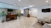 Located at 215 Fairmount Avenue in Lynn, Massachusetts, this single-floor community space is thoughtfully designed with a modern, hospitality-inspired aesthetic. The entrance welcomes visitors through sleek black-framed glass doors, opening into a bright, open lobby finished with large-format porcelain tile flooring that flows seamlessly throughout the entire space.

A dramatic full-height glass partition elegantly separates the entry lobby from the main lounge area, creating a sense of openness while defining distinct zones. The lounge centers around a striking stone-clad feature wall housing a linear electric fireplace beneath a wall-mounted television, serving as the focal point of the room. A generously sized sectional sofa and wooden coffee table set atop a patterned area rug create an inviting gathering space.

Adjacent to the lounge, a stylish wet bar with a quartz countertop and bar seating for multiple guests is illuminated by industrial-style pendant lighting, adding warmth and character. The bar area is framed by wood-toned cabinetry that complements the neutral gray and taupe color palette found throughout.

A dedicated seating alcove with a textured accent wall and warm wood credenza provides a quieter retreat, furnished with patterned armchairs and a round concrete-top coffee table. Recessed lighting throughout ensures a well-lit, comfortable ambiance, making this space ideal for socializing and community gatherings.