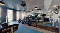 Located at 900 Doverside Dr in Apex, NC, this well-appointed fitness center delivers a comprehensive workout experience within a thoughtfully designed single-floor layout. The main exercise area is spacious and airy, featuring tray ceilings accented with bold teal panels, black pendant lights, and multiple ceiling fans that keep the space comfortable. Large windows along the perimeter flood the floor with natural light while offering views of the surrounding community. Floor-to-ceiling mirrors line the walls, amplifying the sense of space and allowing members to monitor their form.

The cardio zone is generously equipped with a row of Life Fitness ellipticals, treadmills, and stationary bikes, each paired with personal display screens. A dedicated free weights section offers a full dumbbell rack alongside benches for strength training. A Life Fitness functional training cable system anchors the strength area, complemented by multiple selectorized weight machines for a complete resistance workout. Colorful geometric-patterned carpet tiles define distinct workout zones while adding visual energy to the space.

A separate spin studio features dedicated cycling bikes, a large display screen, mirrored walls, and a medicine ball rack for cross-training. A smaller supplementary room with a distinctive mountain-silhouette mural provides a quiet nook with a desk, adding versatility to the overall facility.
