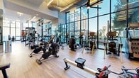 Located at 1401 Union St in San Diego's vibrant Little Italy neighborhood, this single-level fitness facility offers a comprehensive range of indoor and outdoor workout spaces designed for both performance and comfort. The interior gym is thoughtfully divided into distinct training zones. A cardio area features premium equipment including Cascade treadmills, air bikes, ellipticals, and rowing machines arranged across light wood flooring beneath soaring ceilings. Floor-to-ceiling glass walls flood the space with natural light while framing views of the surrounding San Diego skyline. Textured stone-patterned accent walls and large mirrored panels enhance the modern aesthetic throughout. A dedicated strength training zone houses a full dumbbell rack, cable machines, a power rack, and an adjustable bench, complemented by rubber flooring and additional mirrored walls. A Jacob's Ladder stair climber adds further variety to the equipment selection. Outdoors, an expansive turf training area provides space for functional fitness with pull-up rigs, TRX suspension systems, battle ropes, and weight plates. Adjacent to the turf, a stylish lounge area features a pergola structure, wood-paneled walls, mounted displays, lush planters, and modern seating, creating a relaxed outdoor social environment with open city views.