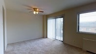 This single-floor space presents a clean, functional layout centered around a carpeted main living area. The room is finished with neutral-toned walls and light beige wall-to-wall carpeting, creating a calm and versatile backdrop suitable for a variety of furniture arrangements. A ceiling fan with an integrated light fixture is centrally mounted, providing both overhead illumination and air circulation throughout the space. A large sliding glass door serves as a prominent architectural feature, fitted with vertical blinds that allow for adjustable light control. The door provides direct access to an outdoor area while framing a view of surrounding greenery, contributing natural light that brightens the interior. Electrical outlets are positioned along the baseboards at functional intervals. The overall ambiance is modest and straightforward, offering a blank canvas that can be personalized to suit various living styles. The single-floor configuration ensures easy accessibility throughout, and the open feel of the main room provides practical versatility for everyday use.