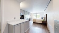 Located at 18 Old Ridgebury Road in Danbury, Connecticut, this efficiently designed studio offers a thoughtfully organized single-floor layout that maximizes every inch of available space. The open-plan living and sleeping area is anchored by warm-toned hardwood-style flooring that runs throughout, creating a cohesive and inviting atmosphere.

The kitchen is fully equipped with stainless steel appliances, including a full-size range, over-the-range microwave, and refrigerator. Gray shaker-style cabinetry extends along the upper wall, providing generous storage, while a white quartz peninsula countertop doubles as a prep and dining surface. A built-in dresser and wall-length desk area offer additional functional storage and workspace alongside the kitchen zone.

The sleeping area is positioned to take full advantage of a large sliding window that frames lush, tree-lined outdoor views, filling the space with natural light. A dark wood accent wall serves as a stylish headboard backdrop, enhancing the room's modern aesthetic.

A dedicated bathroom features tile flooring, a granite-topped vanity, and clean, neutral finishes. The unit also includes an in-unit washer integrated into the kitchen area near the entryway, as well as a closet for additional storage. The overall design balances modern functionality with a calm, neutral palette.