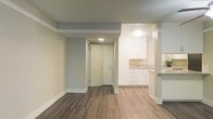 This single-floor space showcases a thoughtfully designed open layout that connects the main living area to the kitchen. The living area features warm-toned, hardwood-style flooring that runs throughout, complemented by soft grey walls and crisp white baseboards, creating a clean and cohesive aesthetic. A textured ceiling adds subtle depth to the space.

The kitchen is visible from the main living area and is outfitted with white shaker-style cabinetry offering generous storage both above and below the counter. Light-toned countertops provide a bright contrast to the flooring, and a raised bar-height counter serves as a partial divider between the kitchen and living space, offering a convenient area for casual dining or workspace.

A paneled entry door is centrally positioned and features a deadbolt lock. Recessed and flush-mount ceiling lighting provides practical illumination throughout both the living and kitchen areas. The overall design balances functionality with a modern, neutral palette, making the space feel clean, well-maintained, and move-in ready.