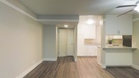 This single-floor space showcases a thoughtfully designed open layout that connects the main living area to the kitchen. The living area features warm-toned, hardwood-style flooring that runs throughout, complemented by soft grey walls and crisp white baseboards, creating a clean and cohesive aesthetic. A textured ceiling adds subtle depth to the space.

The kitchen is visible from the main living area and is outfitted with white shaker-style cabinetry offering generous storage both above and below the counter. Light-toned countertops provide a bright contrast to the flooring, and a raised bar-height counter serves as a partial divider between the kitchen and living space, offering a convenient area for casual dining or workspace.

A paneled entry door is centrally positioned and features a deadbolt lock. Recessed and flush-mount ceiling lighting provides practical illumination throughout both the living and kitchen areas. The overall design balances functionality with a modern, neutral palette, making the space feel clean, well-maintained, and move-in ready.