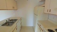 This single-floor space includes a kitchen designed with practicality in mind. The kitchen features light-toned wooden cabinetry lining both sides of the galley-style layout, providing ample storage through upper and lower cabinet units with simple bar-style handles. The countertops complement the cabinetry with a warm, neutral finish. A stainless steel sink is integrated into the counter on one side, while a full-sized white refrigerator anchors the opposite end of the space. A dishwasher is also included beneath the counter, adding convenience. The flooring consists of light-toned tile laid in a classic grid pattern, contributing to the kitchen's clean and bright appearance. The walls are painted in a crisp, neutral white, enhancing the sense of openness within the galley configuration. The layout is efficient, making practical use of the available space by positioning appliances and cabinetry to maximize workflow. Overall, the kitchen presents a straightforward, functional aesthetic with a cohesive neutral palette throughout.