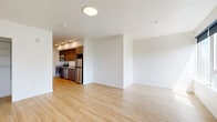 This contemporary studio offers a thoughtfully designed single-floor layout that efficiently combines a kitchen and living area with one full bathroom. Light wood-tone flooring runs throughout the entire space, creating a warm, cohesive aesthetic that complements the clean white walls and modern finishes.

The open-concept kitchen is a standout feature, outfitted with rich wood-grain cabinetry extending to the ceiling for ample storage, white quartz countertops, and a full suite of stainless steel appliances including a refrigerator, electric range, microwave, and dishwasher. Recessed lighting above the kitchen adds a polished touch.

The living area is bathed in natural light from multiple large windows fitted with roller shades, offering views of the surrounding neighborhood and greenery. The generous, unobstructed floor space allows for flexible furniture arrangements. A built-in closet with shelving provides convenient storage.

The bathroom is clean and modern, featuring wood-grain cabinetry with a single vanity sink, a large mirror, and a full soaking tub with shower combo. Wood-look tile flooring ties the bathroom's aesthetic seamlessly to the rest of the unit. The overall ambiance is bright, minimal, and move-in ready.