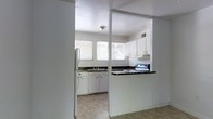 This single-floor space is defined by a practical and straightforward layout centered around a well-appointed kitchen. The kitchen is the focal point of the space, characterized by white cabinetry with black hardware that provides a clean, classic contrast. Dark granite-style countertops run along the perimeter, offering ample prep and workspace. A stainless steel double-basin sink is positioned beneath a wide bank of windows fitted with horizontal blinds, allowing generous natural light to fill the room while maintaining privacy. The kitchen is equipped with a white refrigerator, a dishwasher, and an electric range with an overhead ventilation hood, making it fully functional for everyday use. A half-wall partition with a raised countertop bar separates the kitchen from the adjoining area, creating an open pass-through feel that connects the spaces while maintaining a defined layout. The flooring transitions from light tile in the kitchen to a darker surface in the adjacent area. The white walls and ceilings throughout contribute to a bright, airy atmosphere. The space is compact yet efficiently organized, offering a no-frills environment that is move-in ready and highly functional.