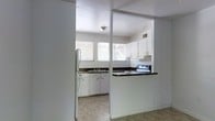 This single-floor space is defined by a practical and straightforward layout centered around a well-appointed kitchen. The kitchen is the focal point of the space, characterized by white cabinetry with black hardware that provides a clean, classic contrast. Dark granite-style countertops run along the perimeter, offering ample prep and workspace. A stainless steel double-basin sink is positioned beneath a wide bank of windows fitted with horizontal blinds, allowing generous natural light to fill the room while maintaining privacy. The kitchen is equipped with a white refrigerator, a dishwasher, and an electric range with an overhead ventilation hood, making it fully functional for everyday use. A half-wall partition with a raised countertop bar separates the kitchen from the adjoining area, creating an open pass-through feel that connects the spaces while maintaining a defined layout. The flooring transitions from light tile in the kitchen to a darker surface in the adjacent area. The white walls and ceilings throughout contribute to a bright, airy atmosphere. The space is compact yet efficiently organized, offering a no-frills environment that is move-in ready and highly functional.